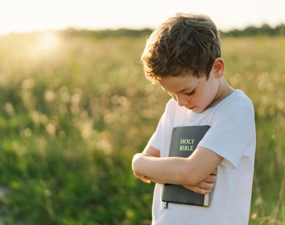 Christian boy holds bible in her hands. Reading the Holy Bible in a field during beautiful sunset. Concept for faith, spirituality and religion. Peace, hope